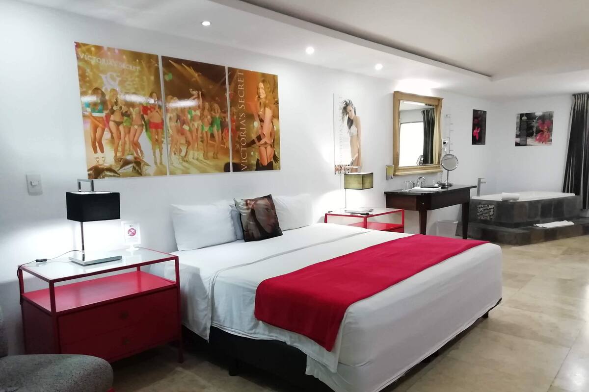 A modern hotel room with a large bed, a bedside table with a lamp, a red nightstand, a small desk, a mirror, a bathroom with a sink and bathtub, and wall art.