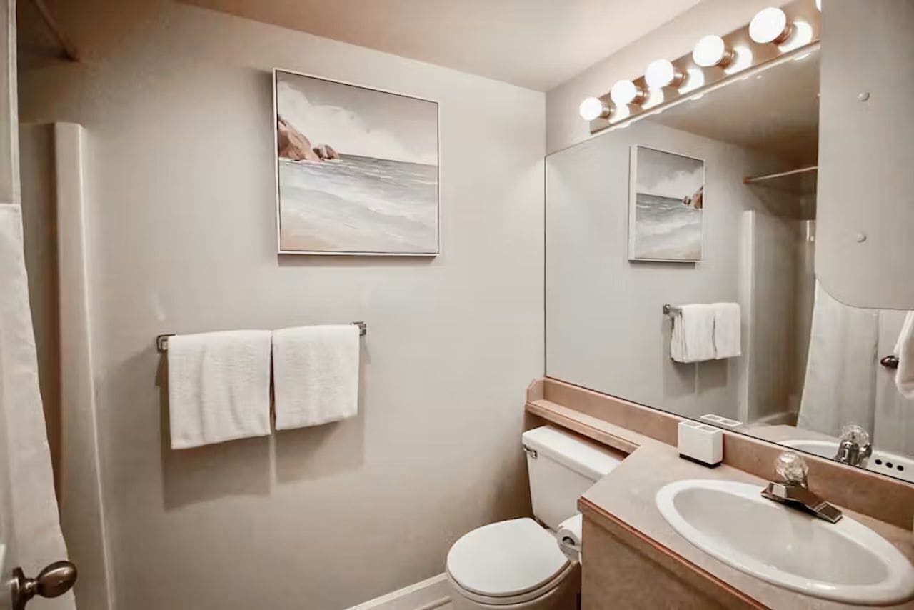 A modern bathroom with a toilet, sink, and a large mirror. There are two towels hanging on the wall and a framed picture above the toilet.