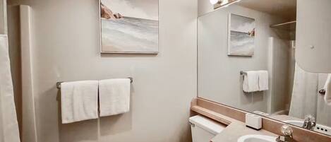 A modern bathroom with a toilet, sink, and a large mirror. There are two towels hanging on the wall and a framed picture above the toilet.