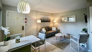 Ein modernes Wohnzimmer mit einer Couch, einem Esstisch und einem Couchtisch. Eine weiße Pendelleuchte, ein Bild an der Wand und eine kleine Pflanze auf dem Beistelltisch sind zu sehen.