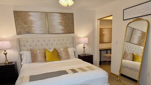 Un dormitorio con una cama grande, dos mesitas de noche, un espejo y una puerta que da a otra habitación.