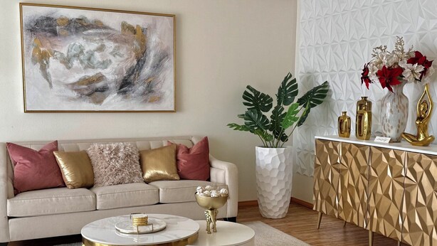 Una sala moderna con un sofá beige, detalles dorados, una mesa redonda de centro, un jarrón blanco con flores rojas y un mueble con estampado geométrico.