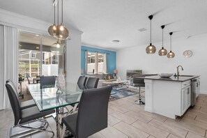 A modern kitchen with a central island, a dining area with a glass table, and a living space with a sofa and TV. - 4 BR Townhome | Free Access to Amazing Resort! (Champions Gate)