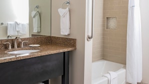 Un baño moderno con encimera de granito, lavabo, espejo y ducha con barra de apoyo.