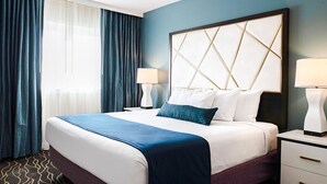 Habitación de hotel con una cama grande, cabecera con diseño geométrico, dos mesitas de noche con lámparas y vistas al exterior a través de cortinas translúcidas.