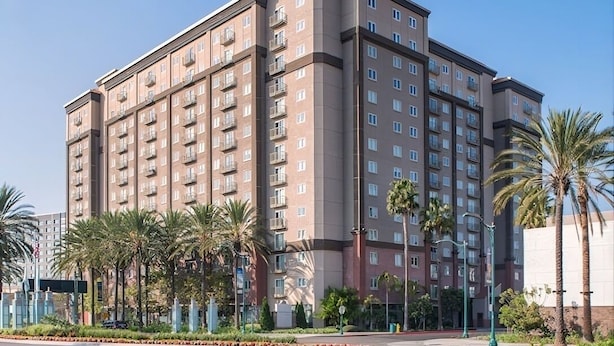 Un hotel de varios pisos con un diseño moderno, con balcones en cada planta y un jardín delantero ajardinado con palmeras.