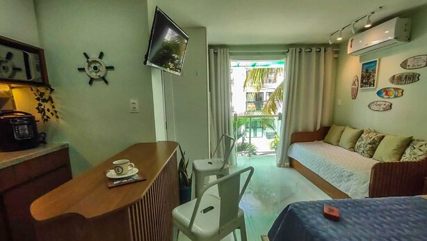 Ett kompakt hotellrum med ett litet kök, en säng, en TV och en balkong med utsikt över palmer.
