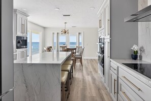 A modern kitchen with a marble island, wooden bar stools, and a view of the ocean.