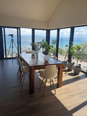 A dining area with a wooden table and chairs, potted plants, and a view of the sea. - Seaview Grove: Wild Atlantic Way Luxury Escape with 180° views over Lough Foyle. (Redcastle)