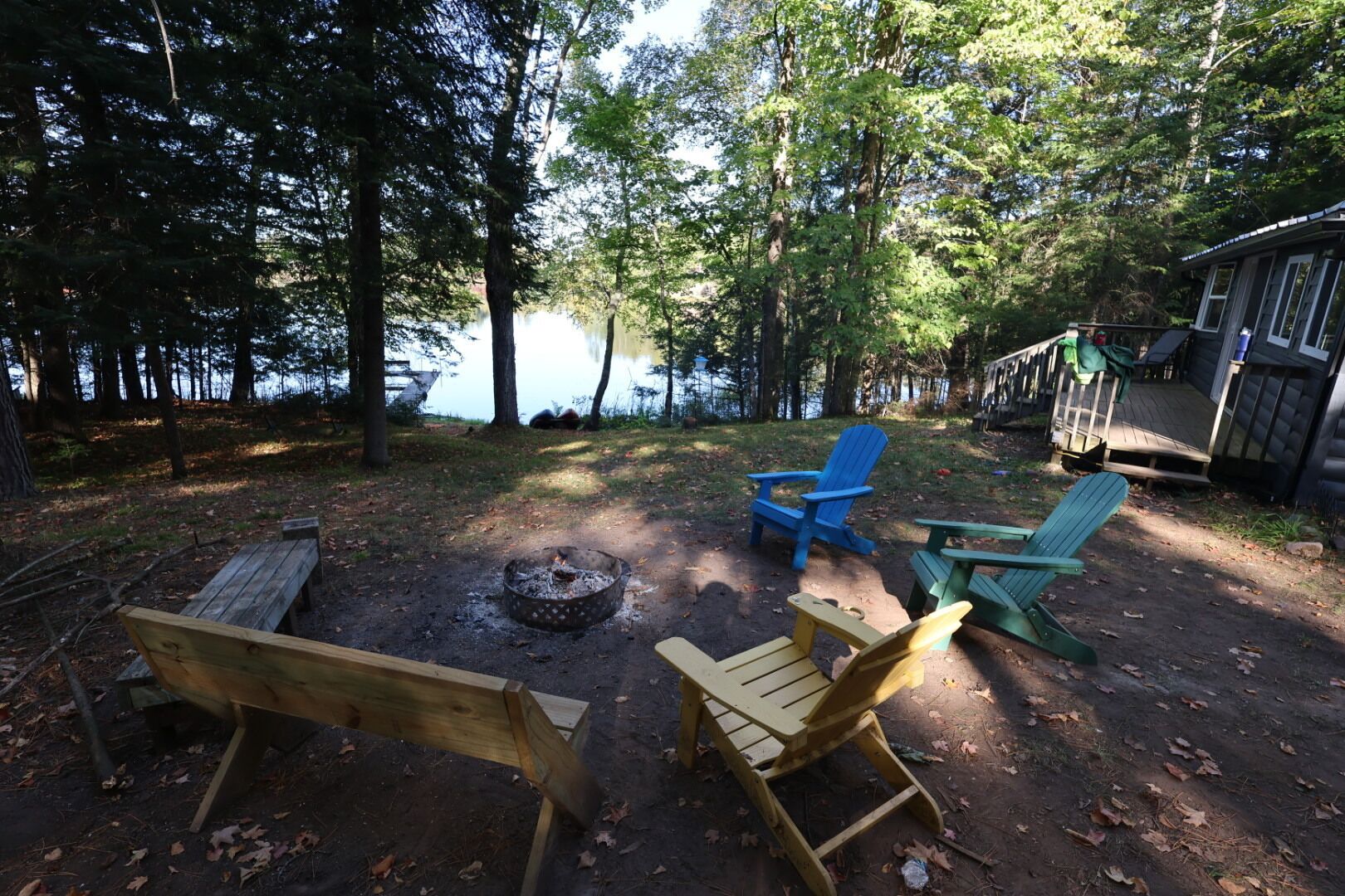 A campsite with a fire pit, wooden benches, and Adirondack chairs by a lake.