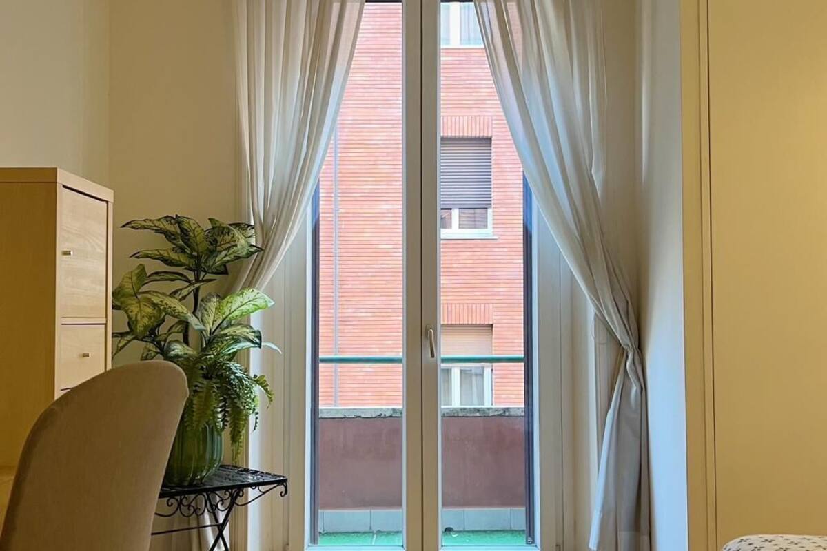 A room with a wooden floor, a chair, a small table with a plant, and a window with curtains.