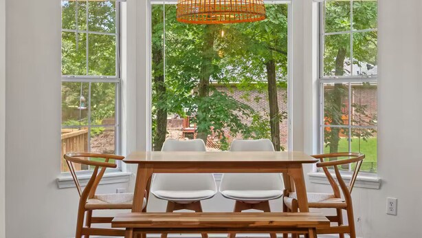 Un coin repas avec une table et des chaises en bois, de grandes fenêtres donnant sur des arbres et un mur en briques, ainsi qu’une suspension orange.