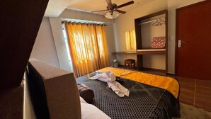 Una habitación de hotel con cama, ventilador de techo, ventana con cortinas, mesita de noche y armario.