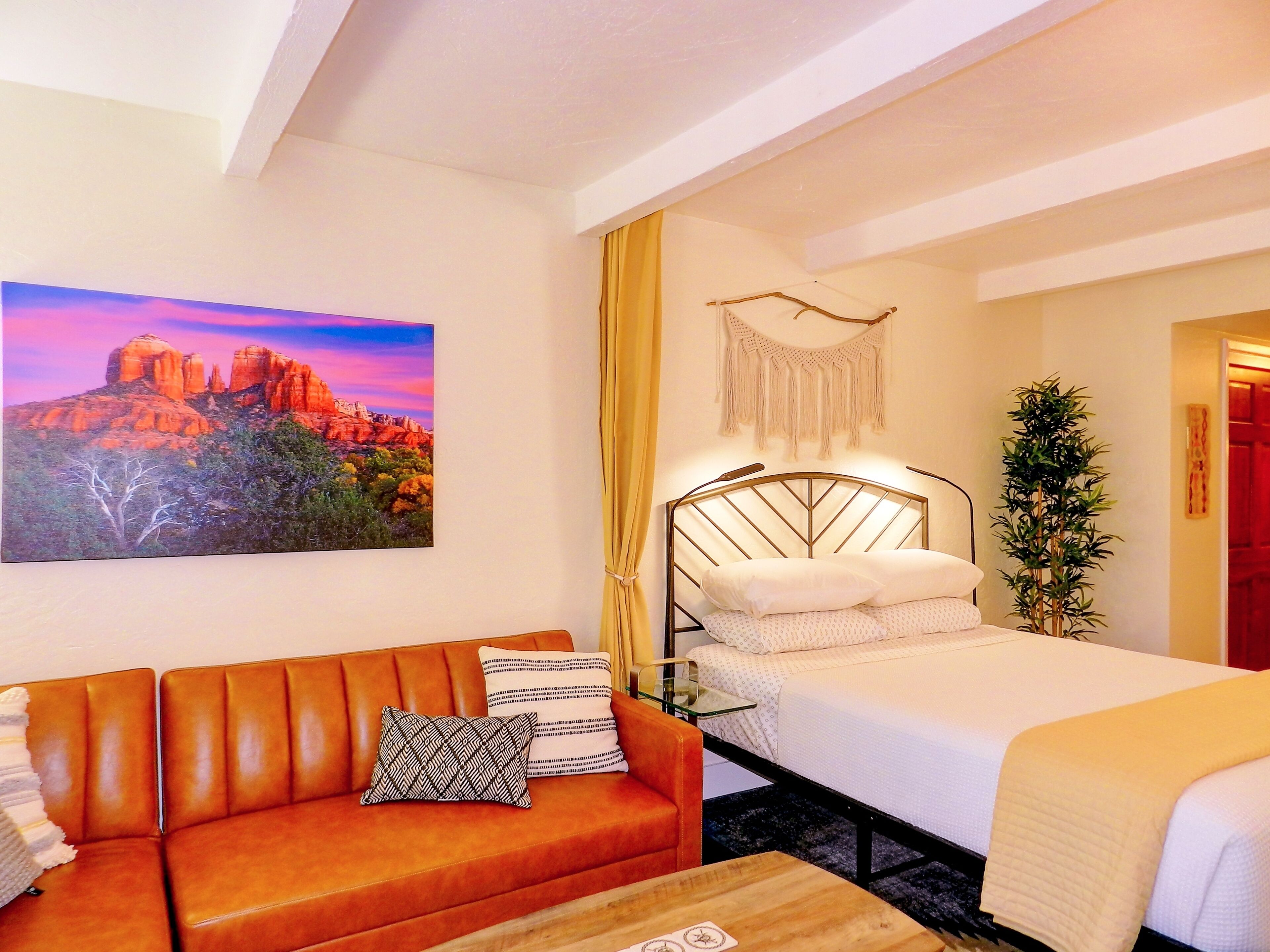 A room with a bed, a couch, a small table, a wall-mounted macrame piece, and a painting of a landscape.