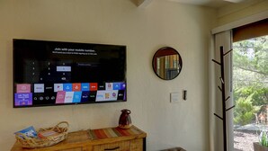 A living room with a flat-screen TV mounted on the wall displaying a mobile number sign-up interface, a wooden console table with a woven basket and a vase, a cushioned bench, and a view of the outdoors through a glass door.