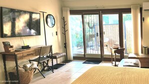 A hotel room with a bed, a desk with a chair, a television, a large window, and a door leading to an outdoor area.