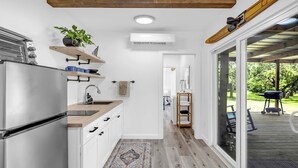 Una cocina moderna con armarios blancos, un refrigerador de acero inoxidable y techo de madera. Hay una planta en un estante, una toalla colgada en la pared y vistas a una terraza con mobiliario exterior.