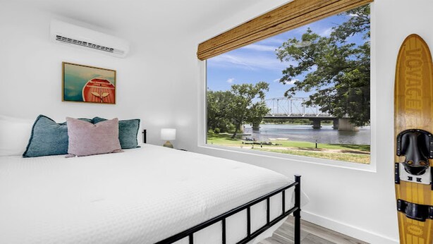 Un dormitorio con un ventanal grande con vistas a un río y un puente, una cama con almohadas, y una tabla de surf apoyada contra la pared.