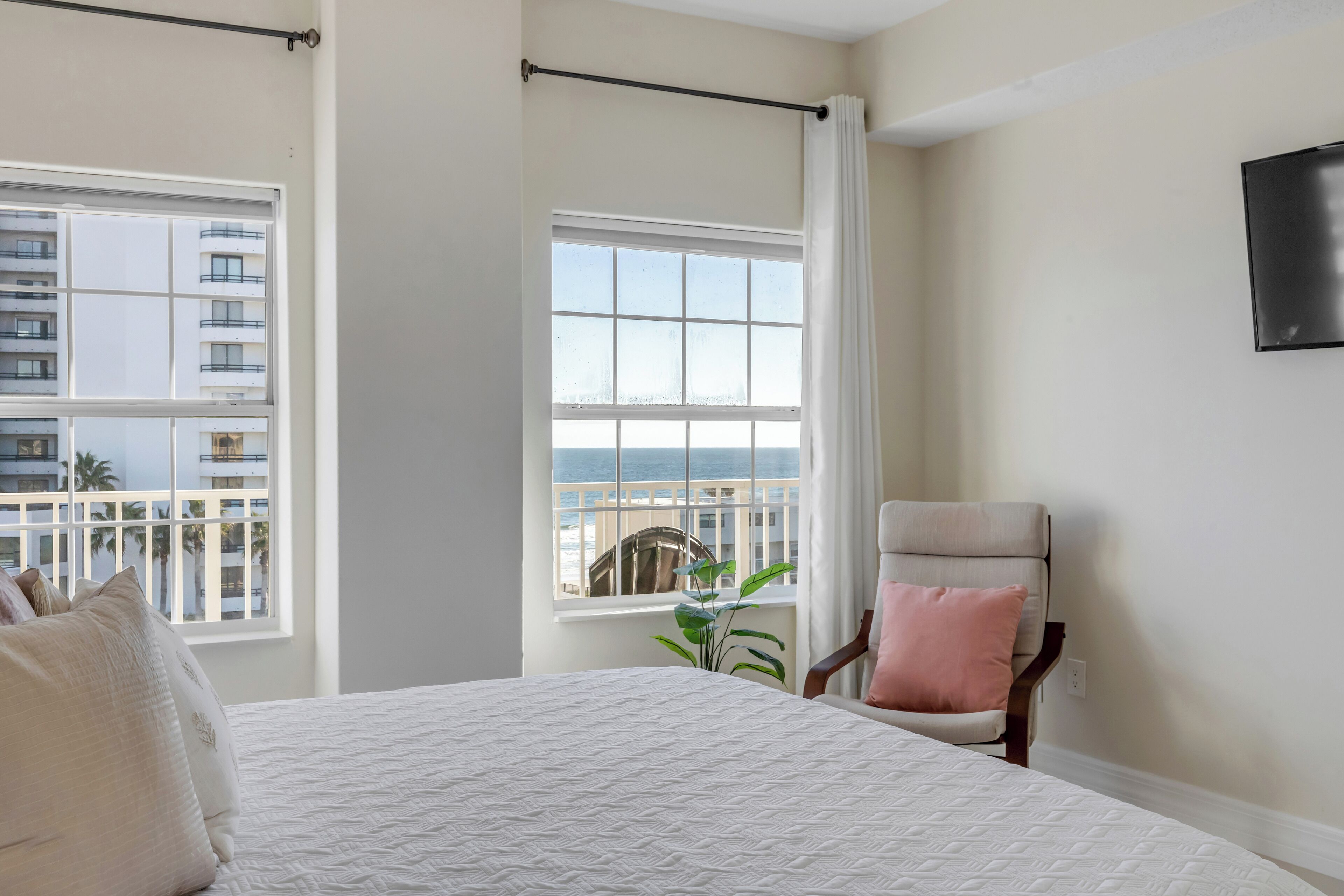 A hotel room with a bed, a chair, a window with curtains, and a view of a building and the sea.