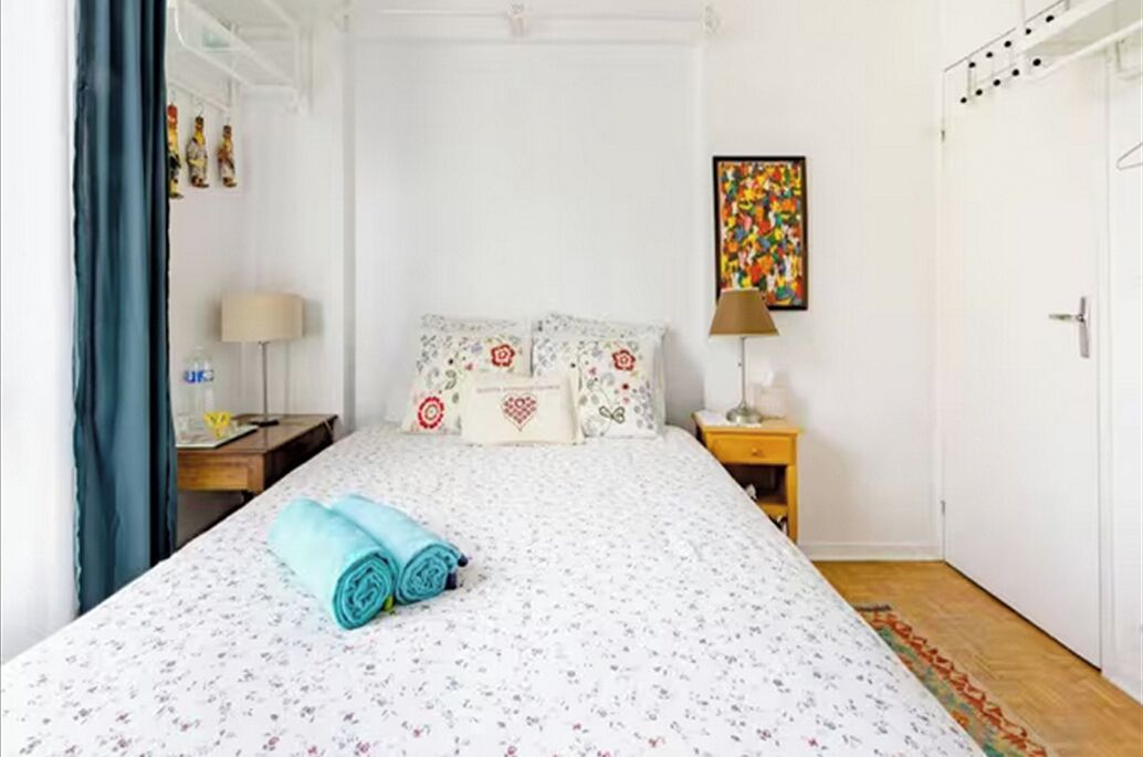 A neatly made bed with floral bedding and rolled towels, a bedside table with a lamp, and a colorful abstract painting on the wall.