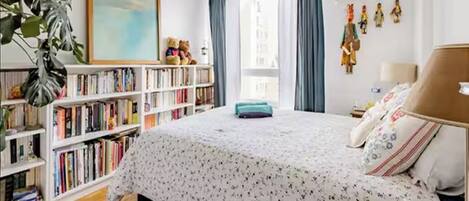 A bedroom with a large bed, a bookshelf, a window with curtains, and a painting on the wall.