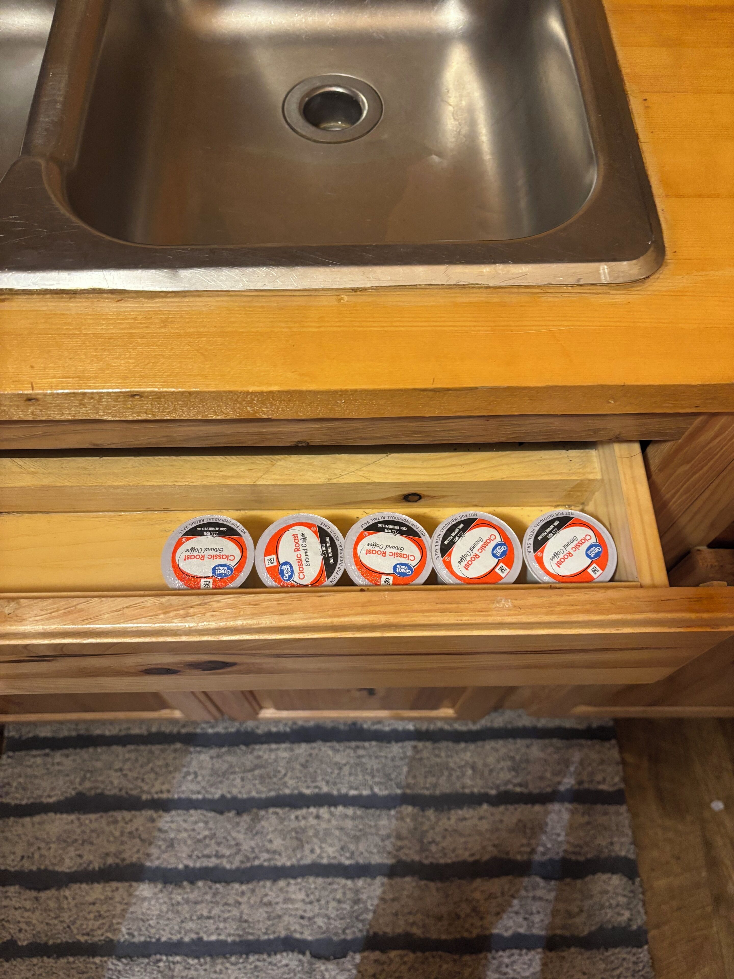 A stainless steel kitchen sink above a wooden drawer containing five containers of Classic Roast coffee.
