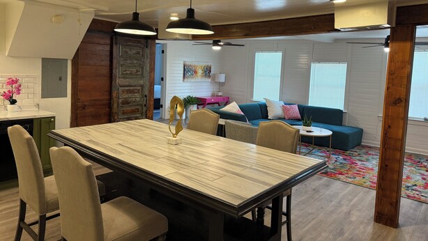 A dining area with a rectangular table, chairs, pendant lights, a kitchen in the background, a living room with a sofa and cushions, and a colorful rug.