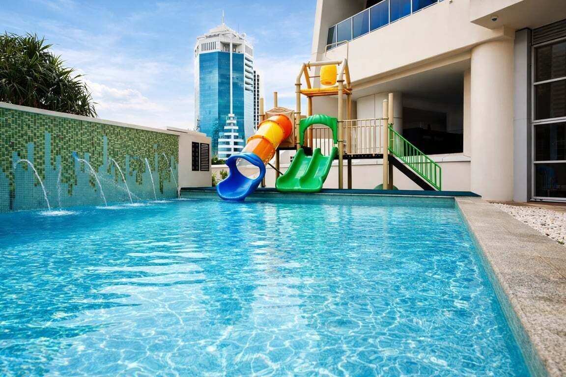 Una piscina con tobogán, un parque infantil y una vista de un edificio alto al fondo.
