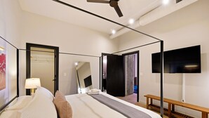 A modern hotel room with a large bed, a bedside lamp, a flat-screen TV mounted on the wall, and a wooden bench. - The Executive | 2BR | Rooftop Pool + Fireplace (St. Louis)