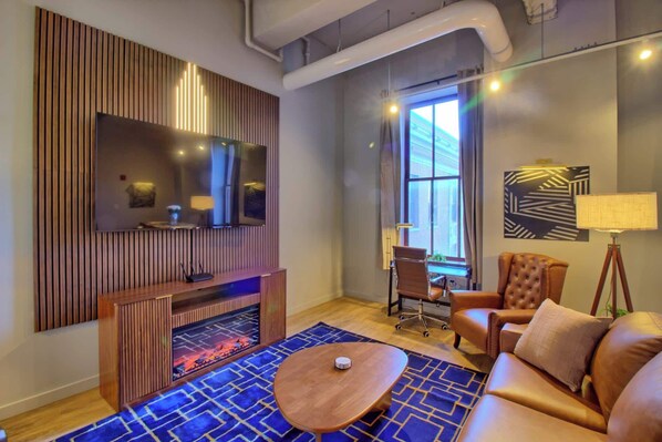 A modern living room with a flat-screen TV, a wooden entertainment unit, a leather sofa, a round coffee table, and a patterned rug. - The Executive | 2BR | Rooftop Pool + Fireplace (St. Louis)
