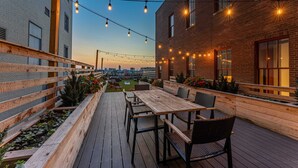 An outdoor patio with wooden tables and chairs, string lights, and a view of the city skyline. - The Executive | 2BR | Rooftop Pool + Fireplace (St. Louis)