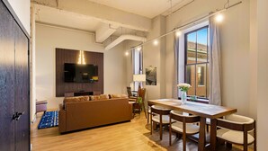 A modern living room with a sofa, dining table, and chairs. There is a TV mounted on the wall, a window with curtains, and a lamp on a side table. - The Executive | 2BR | Rooftop Pool + Fireplace (St. Louis)