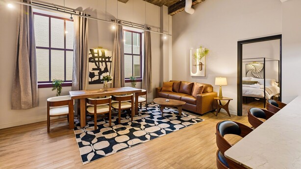 A modern living room with a wooden dining table, a sofa, and a coffee table. There is a large window with curtains, a plant, and a wall-mounted lamp.