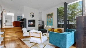 Ein geräumiges Wohnzimmer mit Holzboden, einem weißen Fellstuhl, einem blauen Sofa und einem gelben Sessel. Ein Kamin, ein großes Fenster und ein Gemälde an der Wand sind ebenfalls vorhanden.