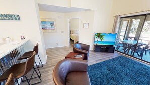 A living room with a TV, a sofa, a coffee table, and a dining area with chairs. - Club Villas Unit 4A (Sunset Beach)