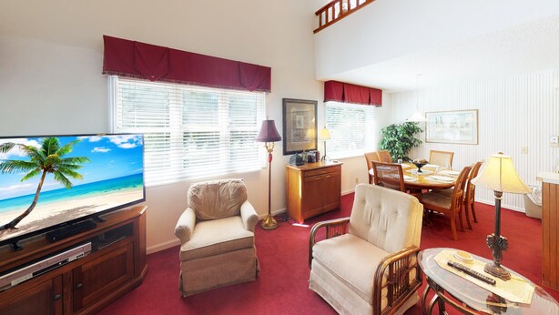 Een woonkamer met een televisie waarop een strand te zien is, een eethoek met een tafel en stoelen, en een rode vloer.