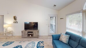 A living room with a flat-screen TV on a wooden stand, a blue sofa, a patterned rug, and a door with blinds. - Club Villas Unit 17D Villa (Sunset Beach)