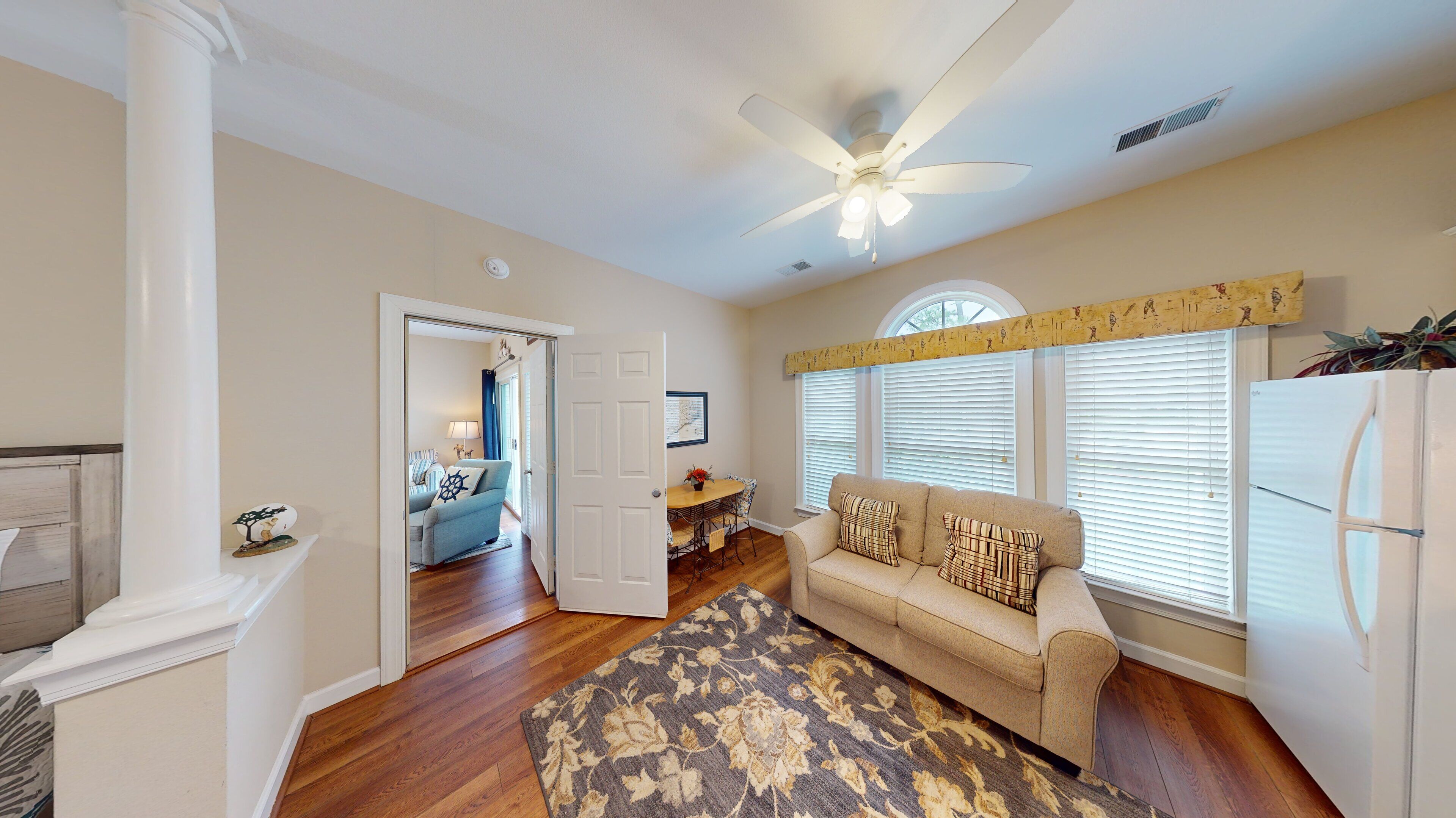 A spacious living room with hardwood floors, a beige sofa, a patterned rug, and a ceiling fan.