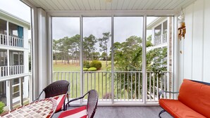 A covered balcony with a view of a golf course, wicker chairs, a red sofa, and a hanging wind chime. - Champions Unit 3707 (Sunset Beach)