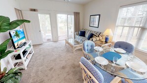 A living room with a television, a sofa, a coffee table, and a dining area with chairs and a table. - Champions Unit 3612 (Sunset Beach)