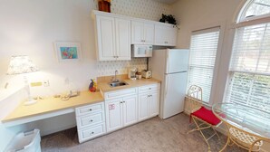 A compact kitchen with white cabinets, a microwave, a toaster, and a sink. There's a small trash bin and a chair with a red cushion. - Champions Unit 3612 (Sunset Beach)