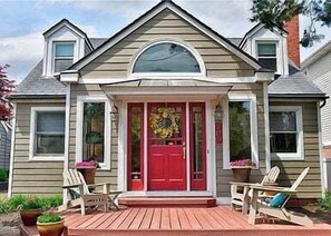 A two-story house with a red front door, a porch with two chairs, and a decorative wreath. - Charming 3 BR North End Beach Cottage One Block to the Beach Private Parking Private Backyard 1478 (Virginia Beach)