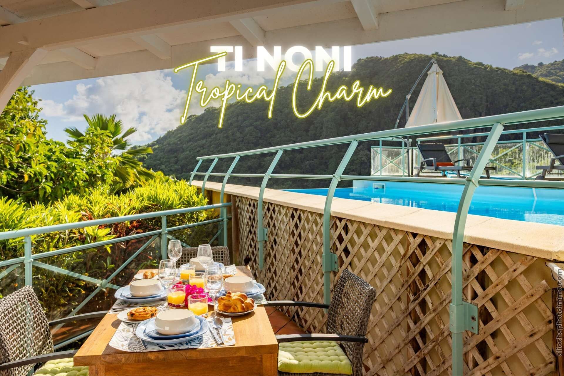 A dining area with a pool view, a table set with plates, glasses, and a bottle, and a sign reading 'TI NONI Tropical Charm'.