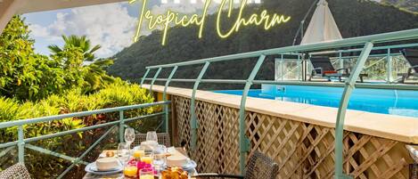 A dining area with a pool view, a table set with plates, glasses, and a bottle, and a sign reading 'TI NONI Tropical Charm'.