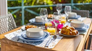 Une table de petit-déjeuner dressée avec des assiettes, des bols, des verres et des viennoiseries, avec en arrière-plan une vue sur la verdure.
