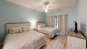 A bedroom with two beds, a ceiling fan, a TV, and a window with curtains. - Club Villas Unit 17D - Lower Level (Sunset Beach)