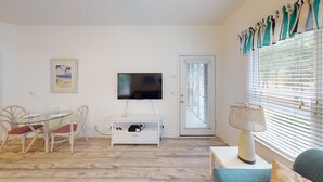 A room with a dining table, chairs, a TV mounted on the wall, a small cabinet, and a window with blinds. - Club Villas Unit 17D - Lower Level (Sunset Beach)