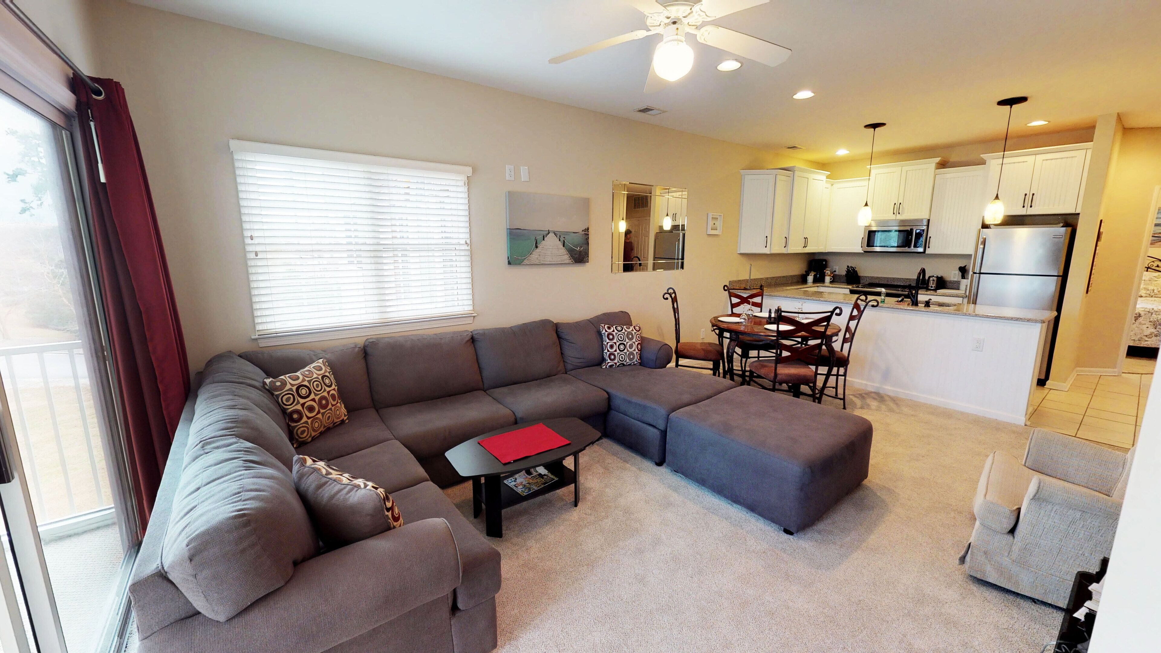 A modern living room with a sectional sofa, a dining table with chairs, and a kitchen area with white cabinets and appliances.