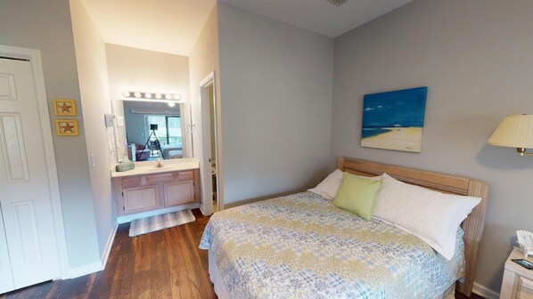 A bedroom with a bed, a nightstand, a lamp, a painting, and a bathroom with a vanity and mirror. - Club Villas Unit 24B (Sunset Beach)
