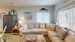 A living room with a beige sectional sofa, a wicker coffee table, a clock, a map on the wall, and a window with blinds. - Champions Unit 3509 (Sunset Beach)